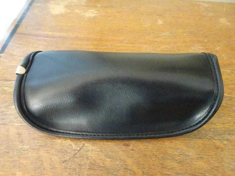 Puede incluir: Un estuche para gafas negro de forma semiovalada con una superficie lisa y mate y bordes cosidos. El estuche presenta un cierre de solapa curvado con un peque&ntilde;o detalle met&aacute;lico de color plateado visible en el lado izquierdo del marco.