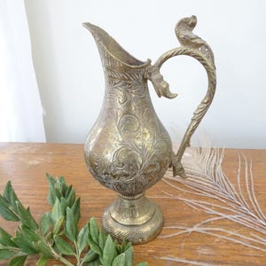 May include: A vintage brass pitcher with intricate floral engravings. The pitcher features a curved handle with decorative elements and a wide spout. The brass has a warm, aged patina, and the pitcher sits on a pedestal base. A decorative item for home decor.