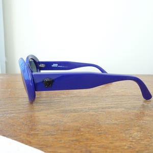 Puede incluir: Gafas de sol azul intenso con monturas gruesas y patillas anchas. Un emblema de cabeza de Medusa negra se muestra en el lateral de la patilla. El interior de la montura incluye el texto GIANNI VERSACE MADE IN ITALY en letras blancas.