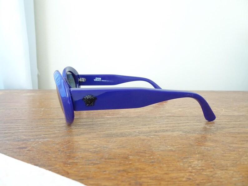 Puede incluir: Gafas de sol azul intenso con monturas gruesas y patillas anchas. Un emblema de cabeza de Medusa negra se muestra en el lateral de la patilla. El interior de la montura incluye el texto GIANNI VERSACE MADE IN ITALY en letras blancas.