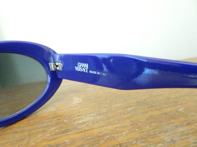 Puede incluir: Un par de gafas de sol azul real con monturas ovaladas. La patilla interior presenta el texto impreso en blanco GIANNI VERSACE MADE IN ITALY. Las gafas tienen un acabado brillante y una bisagra met&aacute;lica que une la montura a la patilla.