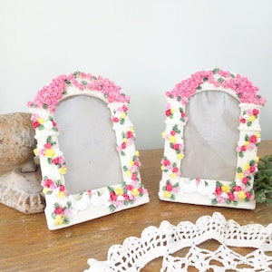 May include: Two white picture frames with arched tops, adorned with pink, yellow, and red roses. The frames have a decorative border and small white doves at the base. The frames are on a wooden surface.