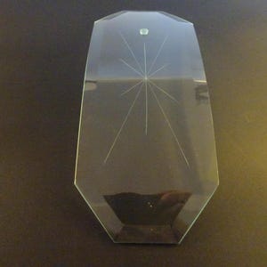 May include: A clear, faceted glass object with a starburst design etched into its surface. The glass has a rectangular shape with a rounded top and a small, circular detail near the top. The object is likely a decorative piece.