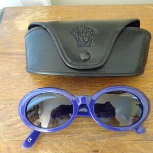 Puede incluir: Gafas de sol ovaladas con montura azul oscuro brillante y lentes oscuras, acompa&ntilde;adas de un estuche protector de cuero sint&eacute;tico negro con un emblema de cabeza de Medusa en relieve en la solapa frontal y un cierre de bot&oacute;n a presi&oacute;n negro.