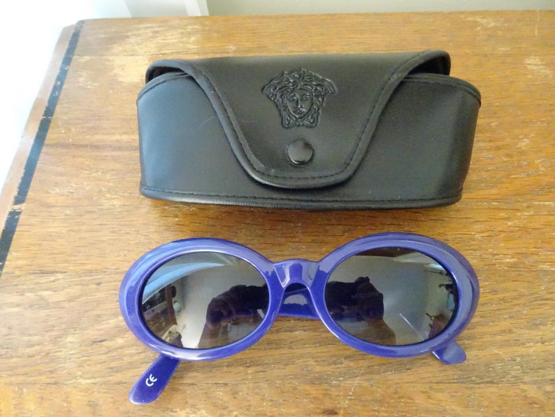 Puede incluir: Gafas de sol ovaladas con montura azul oscuro brillante y lentes oscuras, acompa&ntilde;adas de un estuche protector de cuero sint&eacute;tico negro con un emblema de cabeza de Medusa en relieve en la solapa frontal y un cierre de bot&oacute;n a presi&oacute;n negro.