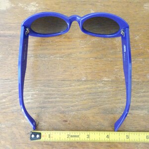 Puede incluir: Gafas de sol azules con lentes ovaladas de tono oscuro y monturas gruesas. Una cinta m&eacute;trica amarilla est&aacute; colocada debajo de las gafas, mostrando una anchura de aproximadamente 14 cm desde el borde exterior de una patilla a la otra.