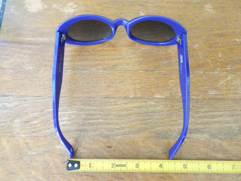 Puede incluir: Gafas de sol azules con lentes ovaladas de tono oscuro y monturas gruesas. Una cinta m&eacute;trica amarilla est&aacute; colocada debajo de las gafas, mostrando una anchura de aproximadamente 14 cm desde el borde exterior de una patilla a la otra.