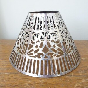 May include: A silver-coloured, cone-shaped lampshade with intricate cut-out floral and vertical bar designs. The lampshade is made of metal and sits on a wooden surface. The interior is black.