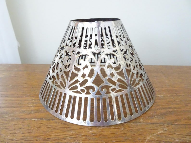 May include: A silver-coloured, cone-shaped lampshade with intricate cut-out floral and vertical bar designs. The lampshade is made of metal and sits on a wooden surface. The interior is black.