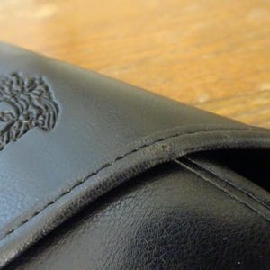 Puede incluir: Una cartera de cuero negro con un emblema de cabeza de Medusa en relieve en la solapa frontal. La cartera tiene costuras tonales visibles a lo largo de los bordes y un acabado mate suave en la superficie exterior, mostrando los rasgos faciales.