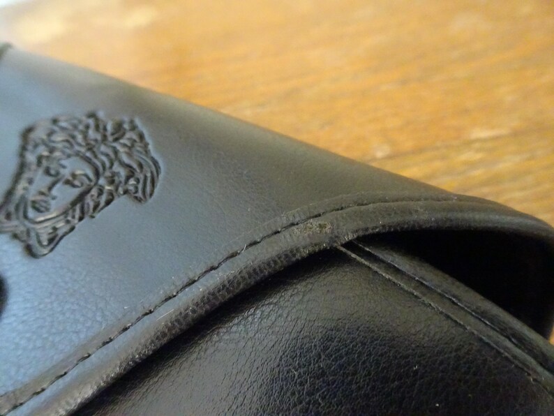 Puede incluir: Una cartera de cuero negro con un emblema de cabeza de Medusa en relieve en la solapa frontal. La cartera tiene costuras tonales visibles a lo largo de los bordes y un acabado mate suave en la superficie exterior, mostrando los rasgos faciales.