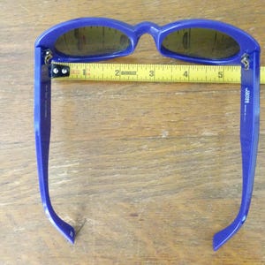 Puede incluir: Gafas de sol azules con lentes oscuras colocadas sobre una cinta m&eacute;trica amarilla. La cinta muestra que el ancho del marco es de aproximadamente 14 cm. La patilla interior presenta el texto 'vogue' y 'made in italy'.