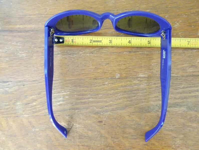 Puede incluir: Gafas de sol azules con lentes oscuras colocadas sobre una cinta m&eacute;trica amarilla. La cinta muestra que el ancho del marco es de aproximadamente 14 cm. La patilla interior presenta el texto 'vogue' y 'made in italy'.