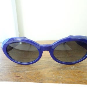 Puede incluir: Un par de gafas de sol con una montura de pl&aacute;stico azul profundo brillante y lentes oscuras de forma ovalada. La montura presenta un dise&ntilde;o grueso y redondeado que rodea las lentes, creando un aspecto llamativo. El puente est&aacute; integrado.