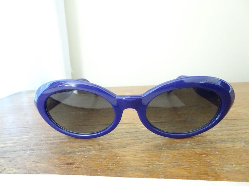 Puede incluir: Un par de gafas de sol con una montura de pl&aacute;stico azul profundo brillante y lentes oscuras de forma ovalada. La montura presenta un dise&ntilde;o grueso y redondeado que rodea las lentes, creando un aspecto llamativo. El puente est&aacute; integrado.