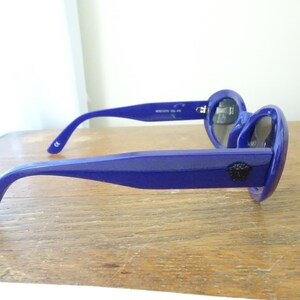 Puede incluir: Un par de gafas de sol de color azul real brillante con monturas gruesas y lentes oscuras. Las patillas son anchas y muestran un peque&ntilde;o emblema circular negro con la cabeza de Medusa en el lateral, cerca de la bisagra.