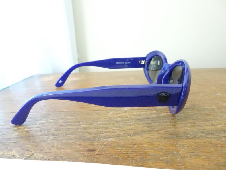 Puede incluir: Un par de gafas de sol de color azul real brillante con monturas gruesas y lentes oscuras. Las patillas son anchas y muestran un peque&ntilde;o emblema circular negro con la cabeza de Medusa en el lateral, cerca de la bisagra.