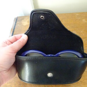 Puede incluir: Un estuche para gafas negro de material similar al cuero con cierre de bot&oacute;n a presi&oacute;n y forro interior negro. La palabra VERSACE est&aacute; grabada en la solapa interior. Una montura de gafas azul oscuro es visible dentro del estuche abierto.