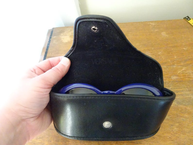 Puede incluir: Un estuche para gafas negro de material similar al cuero con cierre de bot&oacute;n a presi&oacute;n y forro interior negro. La palabra VERSACE est&aacute; grabada en la solapa interior. Una montura de gafas azul oscuro es visible dentro del estuche abierto.
