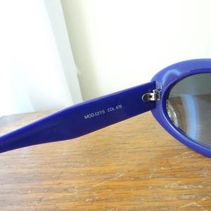 Puede incluir: Un par de gafas de sol azules con monturas ovaladas y lentes oscuras. El interior de la patilla presenta el texto impreso MOD.527/5 COL 476. Las gafas tienen un acabado brillante y una bisagra met&aacute;lica que une la montura con la patilla.