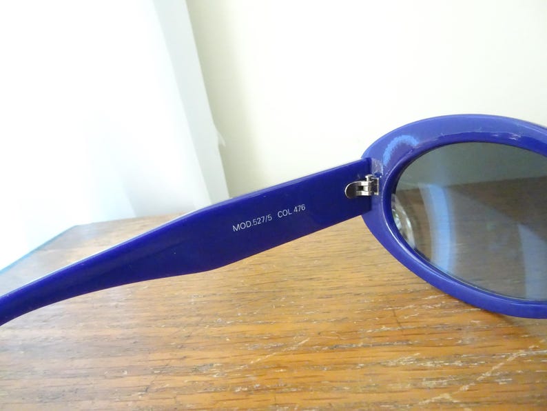 Puede incluir: Un par de gafas de sol azules con monturas ovaladas y lentes oscuras. El interior de la patilla presenta el texto impreso MOD.527/5 COL 476. Las gafas tienen un acabado brillante y una bisagra met&aacute;lica que une la montura con la patilla.