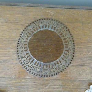 May include: A silver-coloured, openwork metal lampshade ring with a circular design. The ring features a repeating pattern of rectangular and floral shapes. The lampshade ring is on a wooden surface.