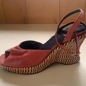 May include: Red leather peep-toe wedge sandal with a woven wedge heel. The sandal features a strap around the ankle and a woven design in red, yellow, and black. The sole is black.