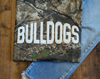 May include: A short-sleeve t-shirt with a camouflage pattern featuring the word BULLDOGS in large white raised lettering across the chest and a pair of light blue denim jeans with a frayed hemline at the bottom of the pant leg.