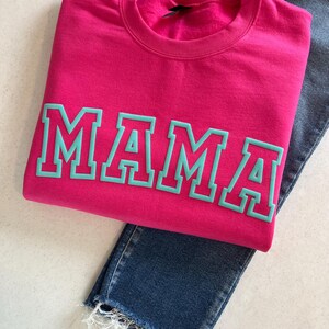 May include: A pink sweatshirt with the word "MAMA" in teal lettering. The letters have a white outline.