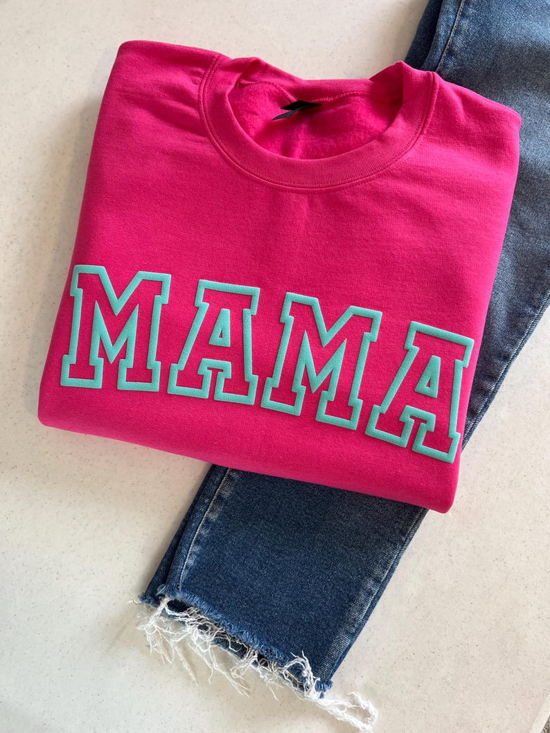 May include: A pink sweatshirt with the word "MAMA" in teal lettering. The letters have a white outline.
