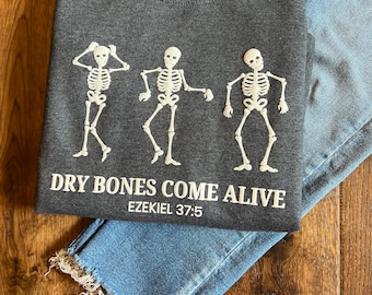 May include: A dark gray crewneck sweatshirt featuring three white skeletons in different dancing poses. Below the skeletons, the text reads "DRY BONES COME ALIVE" in white capital letters, followed by the smaller white text "EZEKIEL 37:5".