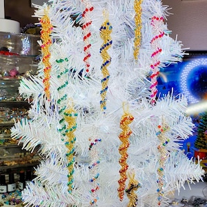 May include: A collection of spiral-shaped beaded icicle ornaments hanging from a white artificial Christmas tree. The ornaments feature various color combinations including gold, red, blue, green, and multi-colored glass beads on wire frames.