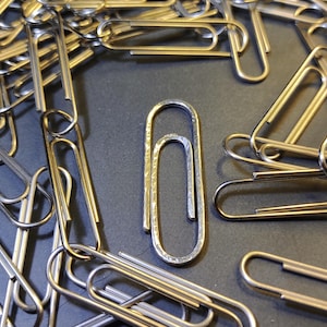 Puede incluir: Una colección de clips de papel plateados esparcidos sobre una superficie oscura. Un clip de papel se alza en el centro, rodeado por otros. Los clips de papel son de metal y tienen un acabado brillante.