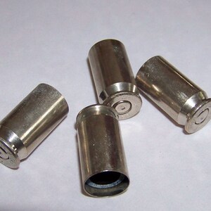 May include: Four metallic, silver-colored bullet casings are scattered on a white surface. The casings are cylindrical with a slightly flared base and a small, circular indentation on the bottom. The casings are empty.