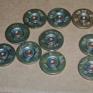 May include: A collection of ten 20 GA shotgun shell casings. The casings are metallic green with a blue ring around the primer and a brass-colored center. The text "WIN HS" and "20 GA" are stamped on the casings.