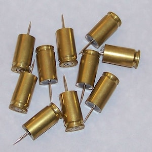 May include: A group of ten brass bullet casings with push pins inserted into the bottom of each casing. The casings are all the same size and shape, and they are arranged in a random pattern on a white surface.