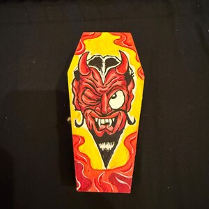 May include: A coffin-shaped wooden box with a devil face design. The devil has red horns, a winking eye, and a goatee. The background is yellow with red flames.