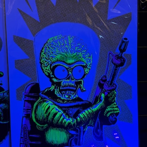 May include: A black and green illustration of a Martian with a ray gun, set against a blue background. The alien has a large head, skull-like face, and is wearing goggles. The artwork is in a comic book style.