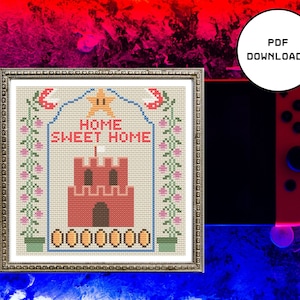 May include: Cross stitch pattern featuring a pixelated castle with the text "Home Sweet Home" above it. The castle is red with white accents and is surrounded by green vines with pink flowers. The pattern is framed in a silver frame.