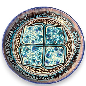 May include: A round ceramic plate featuring a central design divided into four quadrants, each decorated with blue and white floral patterns. The plate has a dark brown and blue border with intricate black scrollwork and a light blue rim.
