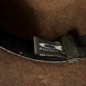 May include: Close-up of a brown hat with a black interior. The hat has a tag that reads "August". The hat appears to be made of a felt-like material.