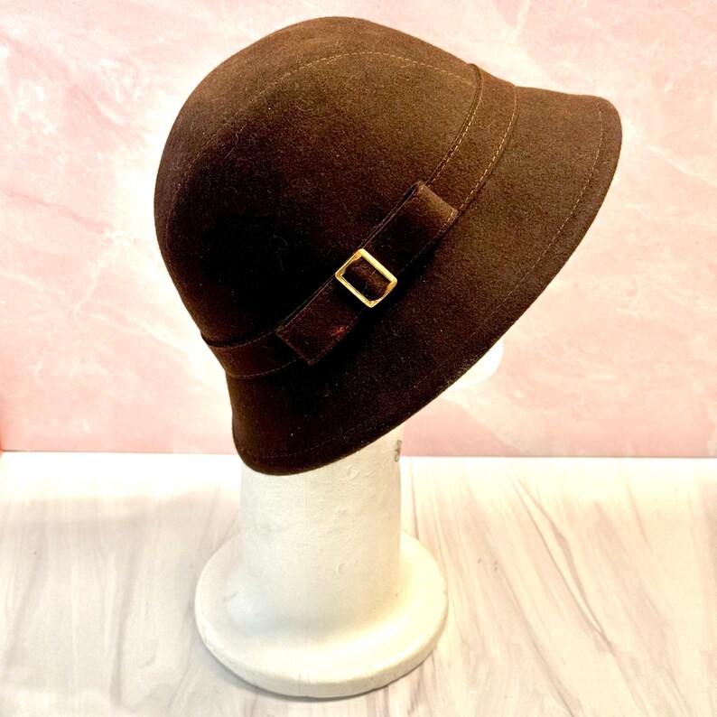 May include: A brown cloche hat with a rounded crown and a small brim. The hat features a decorative band with a bow and a gold-colored buckle. The hat is displayed on a white mannequin head.