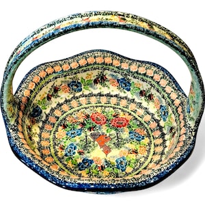 May include: A ceramic basket with a handle, featuring a floral design in red, blue, and peach hues. The rim and handle are detailed with intricate patterns, set against a white background. The basket has a scalloped edge.