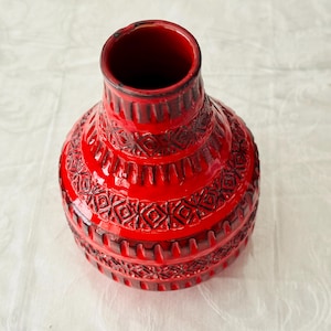 May include: A bright red ceramic vase with a narrow neck and a wide, rounded body. The surface features a repeating dark brown geometric pattern consisting of diamonds, vertical lines, and horizontal bands embossed into the glossy finish.