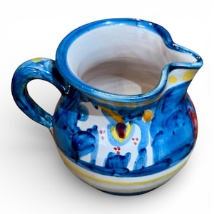 May include: A small, hand-painted ceramic pitcher with a blue and white floral design. The pitcher has a handle and a spout, with yellow and red accents. The rim is blue and the interior is white.