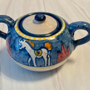 May include: A hand-painted ceramic sugar bowl with a lid and two handles. The bowl is primarily blue with white, yellow, orange, and red accents. The design features a white horse and other abstract shapes.