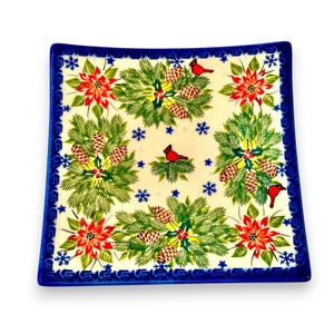 May include: A square ceramic plate with a cream-colored base featuring a festive pattern of red poinsettias, green pine branches, brown pinecones, and red cardinal birds. The design includes small blue snowflakes and a dark blue border.