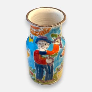 May include: A ceramic vase with a flared rim and a dark brown edge. The exterior features a hand-painted scene on a bright blue background, depicting a person in a hat and red shirt harvesting purple grapes, surrounded by yellow and orange sun motifs.