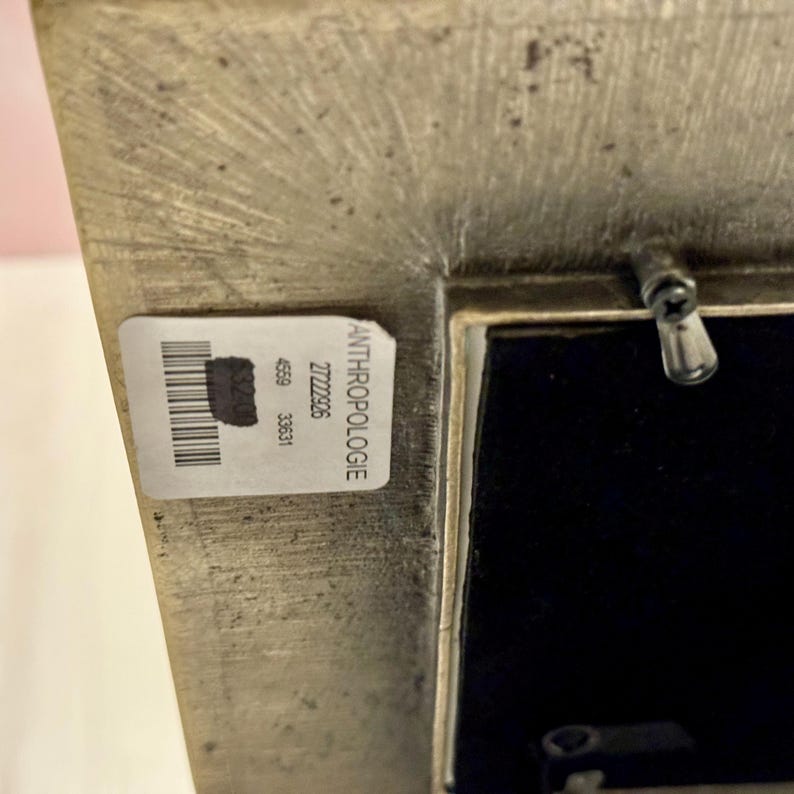 Puede incluir: Un marco de fotos rectangular de tono met&aacute;lico con una superficie texturizada. El marco tiene una etiqueta que dice "ANTHROPOLOGIE" con un c&oacute;digo de barras y otros n&uacute;meros. El interior del marco es negro.