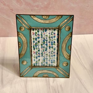 Puede incluir: Un marco de fotos rectangular con acabado esmaltado turquesa y detalles dorados. El marco tiene un inserto con estampado floral. El marco mide aproximadamente 15 cm de alto y 10 cm de ancho.